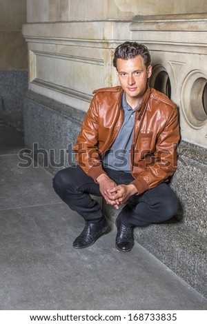 Dressing in a brown leather jacket, gray undershirt, black pants,  a young handsome guy is squatting in the corner of a building, charmingly looking at you. / Portrait of Young Guy