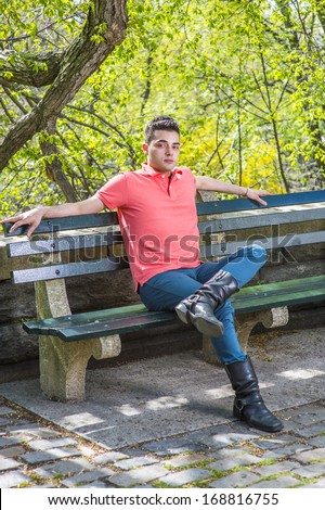 Dressing in a red Polo shirt, green pants and long leather boots, a young handsome guy is sitting on a long chair  in the morning of a spring season, relaxing / Relaxing Outside