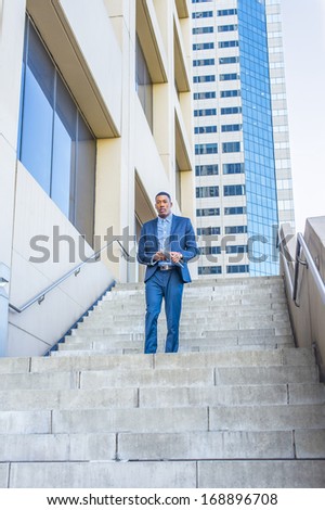 Holding a tablet computer, a young handsome black businessman is confidently walking down stairs outside of a business building. / Portrait of Businessman