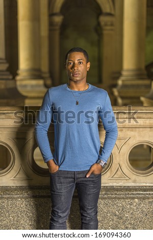 Dressing in a light blue, long sleeve T shirt, hands putting in pockets, a young handsome black student is standing at a hallway and looking at you. / Portrait of Young Black Guy