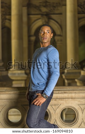 Dressing in a light blue, long sleeve T shirt, a young handsome black student is standing at a hallway, confidently looking forward. / Portrait of Young Black Guy