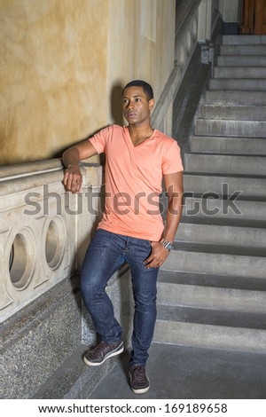 Dressing in a light orange short sleeve V neck shirt, jeans and leather sneakers,  a young handsome black student is standing downstairs by railing into deeply thinking. / Deeply Thinking