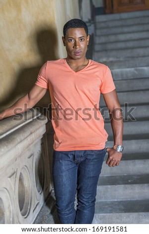 Dressing in a light orange short sleeve V neck shirt, jeans,  a young handsome black student is standing downstairs by railing, charmingly looking at you. / Portrait of Young Student