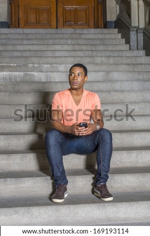 Dressing in a light orange short sleeve V neck shirt, jeans, a young handsome black student is sitting on stairs outside an office building, checking messages on his mobile phone. / Text