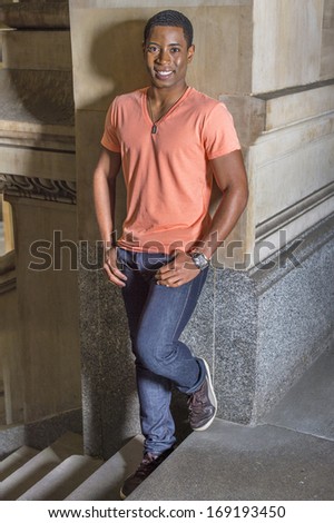 Dressing in a light orange short sleeve V neck shirt, jeans and leather sneakers,  a young handsome black student is leaning against a wall by stairs, smilingly looking at you. / Happy Black Guy