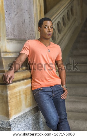 Dressing in a light orange short sleeve V neck shirt, jeans,  a young handsome black student is standing downstairs against a column, charmingly looking at you. / Portrait of Young Black Guy