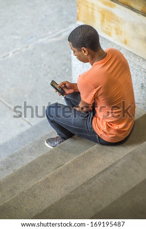 A young handsome black guy is sitting on stairs on the corner, checking messages on his mobile phone. / Text Anywhere