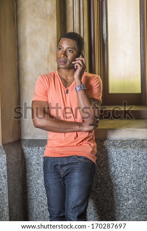 Dressing in a light orange short sleeve V neck shirt, jeans,  a young handsome black student is standing by a small window, listening to the phone. / Making a Phone Call