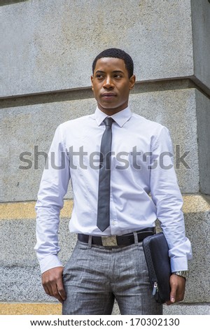 Dressing in white shirt, a black tie, gray pants, carrying a briefcase, a young handsome black college student is standing by a stone wall, confidently looking forward. / Portrait of College Student