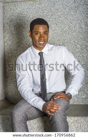 Dressing in white shirt, a black tie, gray pants, wearing a watch, hands resting on his lap, a young black college student is sitting on a stone bench, smilingly looking at you. /College Student