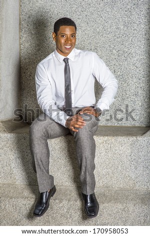 Dressing in white shirt, a black tie, gray pants, leather shoes, wearing a watch, hands resting on his lap, a young black student is sitting on a stone bench, smiling. / Portrait of College Student
