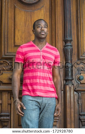 Wearing a red, pink stripe Henley V Neck shirt, gray pants, a bracelet, a young handsome black guy with a little mustache is standing by an old fashion style doorway, looking forward. /Young Black Guy