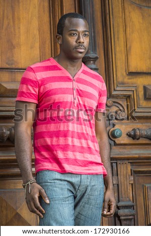 Wearing a red, pink stripe Henley V Neck shirt, gray pants, a bracelet, a young handsome black guy with a little mustache is standing by a old fashion style door, lost in thought. / Young Black Guy