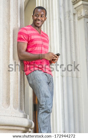 Dressing in red, pink stripe Henley V Neck T shirt,  gray pants, wearing a bracelet, a young black guy is standing by a column, checking messages on his smart phone. / Text