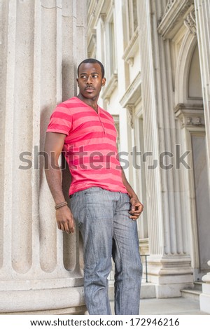 Dressing in red, pink stripe Henley V Neck T shirt,  gray pants, wearing a bracelet, a young black graduate student is standing outside an office building on campus, confidently looking forward.