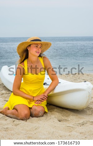 Wearing a bright yellow sundress, a straw hat,  a pretty girl is kneeling down on the beach by a small white lifeguard boat, relaxing. /Beach Vacation