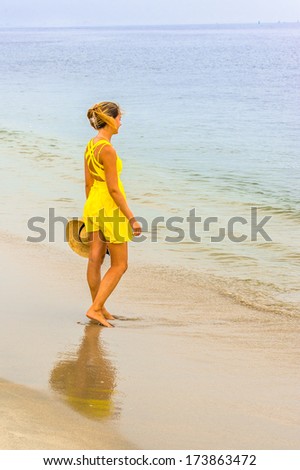 Dressing in a bright yellow sundress, hair clipped on the back of her head, holding a straw hat, a young pretty girl is walking on the beach with barefoot, relaxing. / Walking on Beach