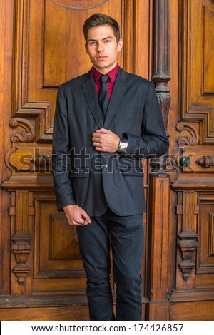 Dressing in a red undershirt, a black blazer, black jeans and a black tie, a young handsome college student is standing in the front of old fashion style door, looking forward. /Young College Student
