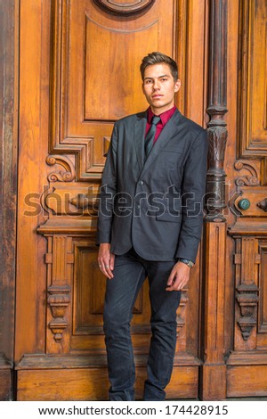 Dressing in a red undershirt, a black blazer,  jeans,  tie,  a handsome college student is standing in the front o old fashion style door, looking at you. / Portrait of Young College Student