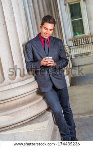 Dressing in a red undershirt, a black blazer,  jeans,  tie, a young handsome college student is leaning against a column outside an office, checking messages on his smart phone. / Text