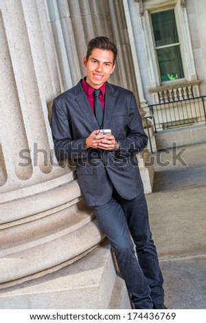 Dressing in a red undershirt, a black blazer,  jeans,  tie, a young handsome college student is leaning against a column outside an office, checking messages on his smart phone. / Text