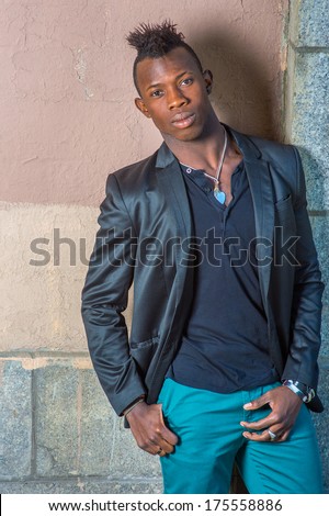 Dressing in a black blazer, green pants, a young black guy with mohawk hair is standing against the wall, his head tilted one side, confidently looking at you. / Portrait of Young Black Guy