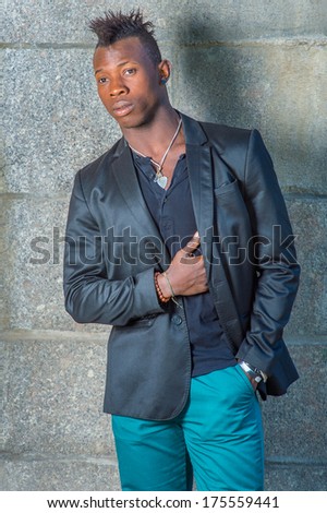 Dressing in a black blazer, green pants,  a hand in his pocket, a hand putting on his chest, a young black guy with mohawk hair is standing against the wall, looking forward. / Portrait of Black Guy