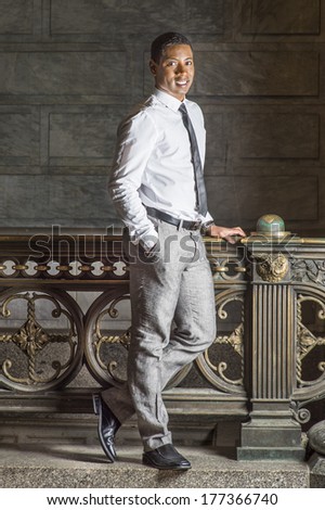 Wearing a white shirt, a black tie, gray pants, a black male college student is standing by a railing on campus, confidently looking at you / Portrait of College Student