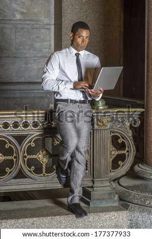 Wearing a white shirt, a black tie, gray pants, a young handsome black college student is standing by a railing on campus, thinking, working on a computer. / Study Anywhere