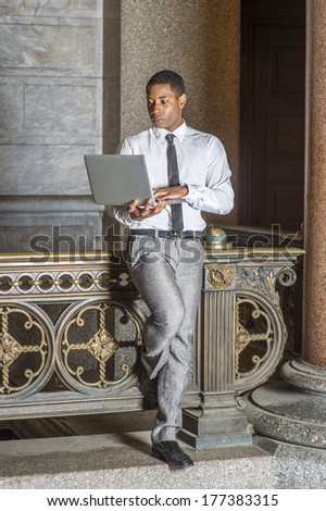 Wearing a white shirt, a black tie, gray pants, a young handsome black college student is standing by a railing in a hallway on a campus, working on a laptop computer. / Study Anywhere