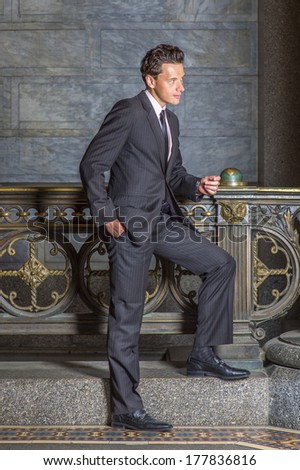 Wearing a black striped jacket, pants, tie, leather shoes, one hand putting in a pocket,  a young businessman is standing by old fashion style railing in a hallway, looking forward and thinking.