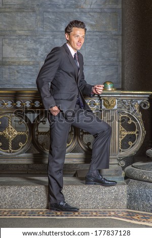 Wearing a black striped jacket, pants, tie, leather shoes, one hand putting in a pocket,  a young businessman is standing by old fashion style railing in a hallway, looking forward and thinking.