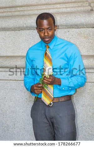 Dressing in a light blue shirt, colorful pattern tie, a young black guy is holding a white rose, standing by a wall, lost in thought. / Thinking about You