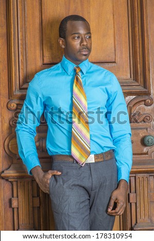 Dressing in a light blue shirt, gray pants, a pattern tie, a hand putting in pocket, a black businessman is standing by a old fashion style door, looking down, lost in thought. / Black Businessman