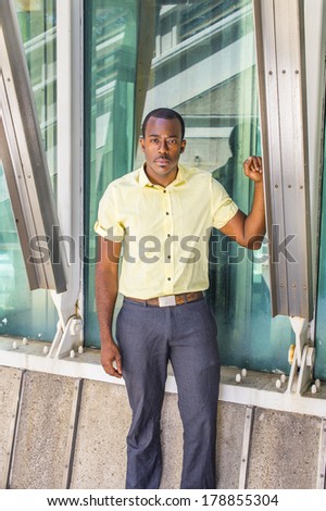Dressing in a light yellow shirt, gray pants, a young attractive black guy is standing by a triangle structure, looking at you. / Portrait of Black Guy