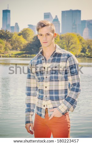 Portrait of Young Man. Dressing in a long sleeve, patterned shirt, red pants, one hand putting in pocket, a young guy standing by a lake. High business buildings in background. Instagram effect.