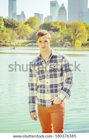 Portrait of Young Man. Dressing in a long sleeve, patterned shirt, red pants, one hand putting in pocket, a young guy is standing by a lake. High business buildings in background. Instagram effect.