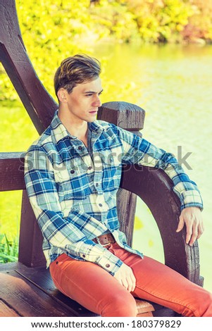 Young man relaxing. Dressing in a long sleeve, patterned shirt, red pants, one young guy is sitting on bench by water, waiting for you. Instagram X-Pro II filter effect.