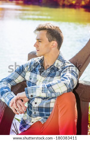 Portrait of Young Handsome Man. Dressing in long sleeve, patterned shirt, red pants, one young guy is causally sitting on bench by water, confidently looking forward. Instagram Toaster filter effect.