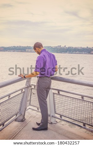 Dressing in a purple shirt, gray pants and leather shoes, a young man with a little beard is facing a river, lowing down his head into deeply thinking. Instagram Sierra effect. / Praying