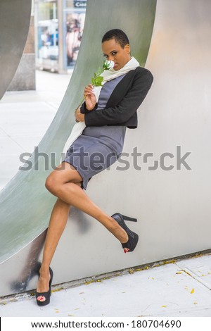 Pretty Black Woman with White Flower. Dressing in woolen cropped jacket, dress, long scarf, high heels, holding white rose,  a young black lady is relying on a metal structure, smelling, thinking.