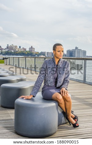 Woman Waiting for You. Dressing in a gray patterned faux fur jacket, a woolen fitted dress, open toes shoes, a professional lady is sitting on a modern style bench on the dock, relaxing, thinking.