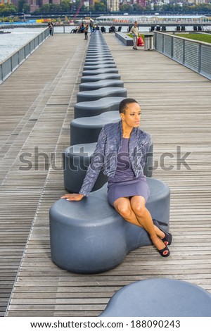 Woman Waiting for You. Dressing in a gray patterned faux fur jacket, a woolen fitted dress, open toes shoes, a young professional lady is sitting on a modern style bench, relaxing, thinking.
