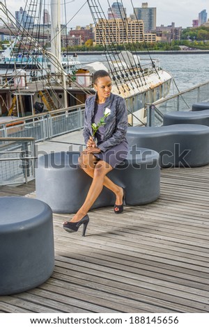 Dressing in faux fur jacket, a woolen fitted dress, open toes shoes, holding a white flower,  a young black woman is sitting on a deck, waiting for you. The background is a harbor.
