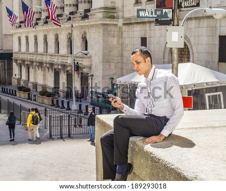 Man Texting on Wall Street.  Wearing a white shirt, black pants, under sunshine,  a young handsome guy is sitting outside office building, texting on his cell phone. Wall Street sign in background.