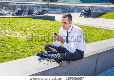 Man Texting Outside. Wearing a white shirt, black pants, leather shoes, sunglasses on the ground, a young guy is checking message on his mobile phone.