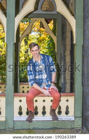 Young Man Relaxing Outside. Dressing in a blue and white pattern shirt,  a blue hood vest,  red jeans and brown leather boot shoes, a young guy is sitting by a pavilion, waiting for you.