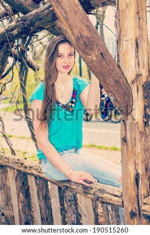 Portrait of Teenager.  Dressing in a blue sleeveless top, fashionable jeans, a beautiful teenage girl is sitting on a wooden fence, charmingly looking at you. Instagram Nashville effect.