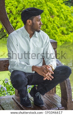 Rainy Day, a little foggy, wet feel. Dressing in a long sleeves shirt, a ivy cap,  a young handsome black guy is squatting by a lake in a raining, foggy day, looking away and thinking.