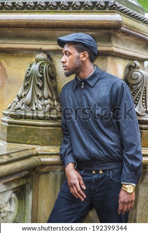 In the rain, a foggy, wet, blur feel. Wearing a waterproof jacket, a ivy cap,  a young black man is standing outside in the corner, waiting, looking around.